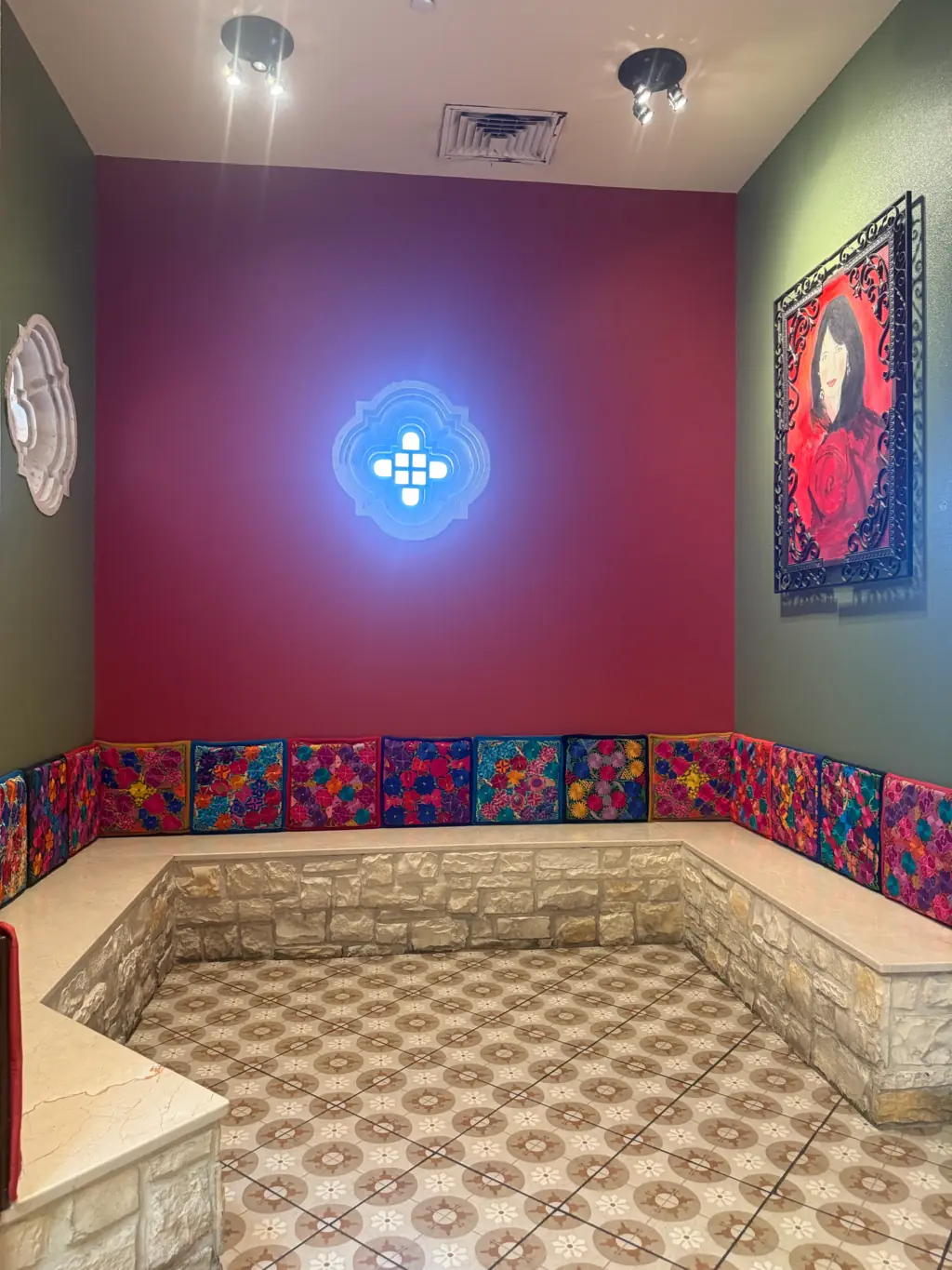 El rincón de la pintura en Sandra's Cuisine: un medallón iluminado en la pared roja, la pintura de la portada en su marco de hierro forjado, una franja de azulejo de colores y una banca de cantera