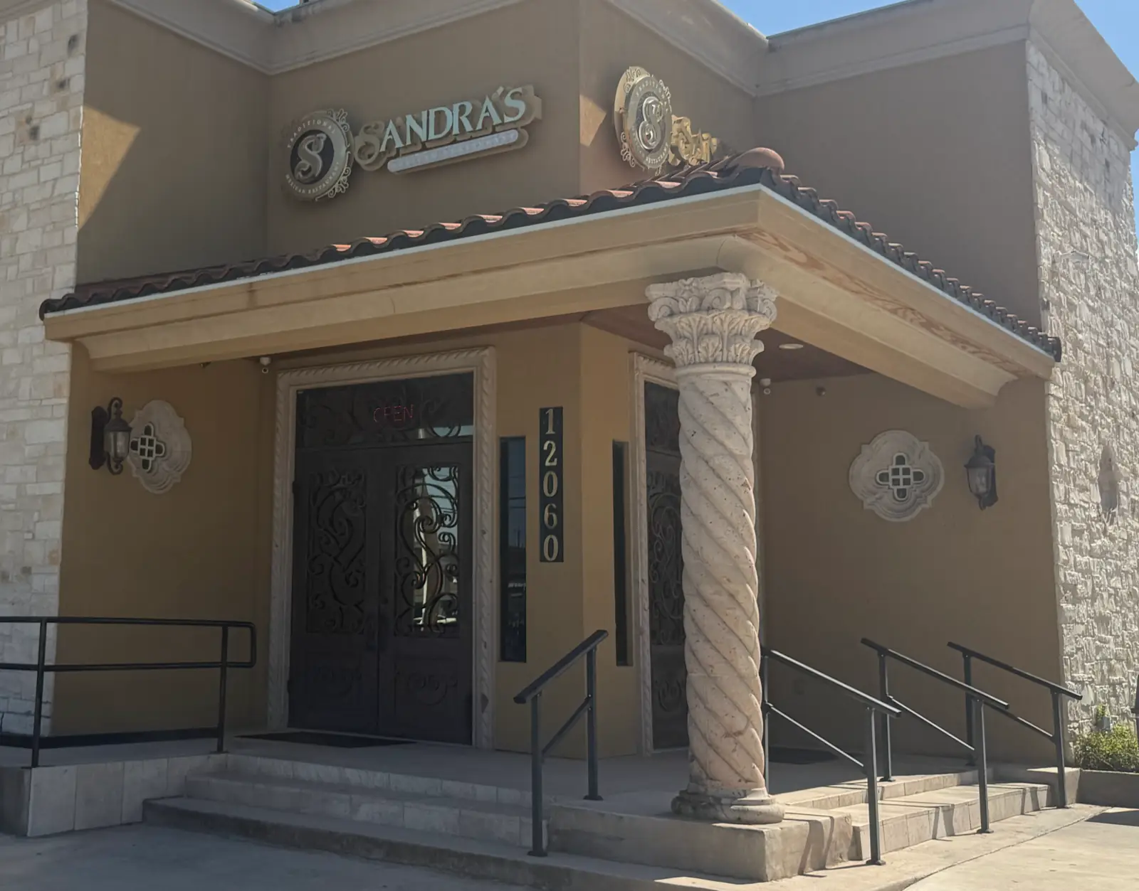 La entrada de Sandra's Cuisine en 12060 Potranco Rd: puertas dobles de hierro y vidrio, una columna salomónica de cantera y el tejado de teja del pórtico